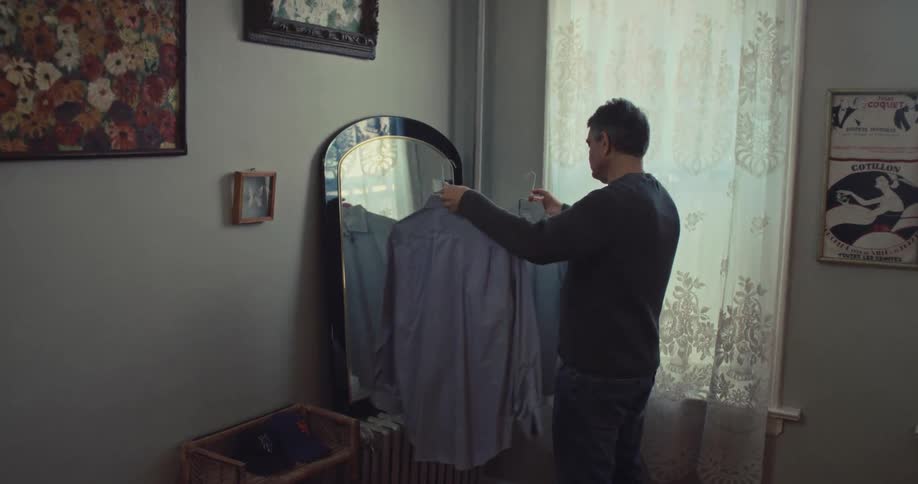 A man stands in a softly lit bedroom beside a tall mirror and lacecurtained window holding up 2 buttondown shirts on a hanger and studying it carefully as if deciding whether to wear it