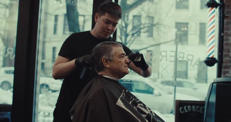A young barber wearing black gloves uses a comb and clippers to trim the hair of an older man sitting in a chair inside a shop while city streets and parked cars are visible through the large glass window in the background