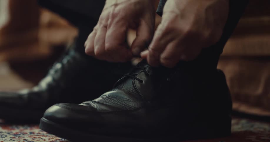 A man ties the laces of a black leather dress shoe while sitting indoors on a patterned rug