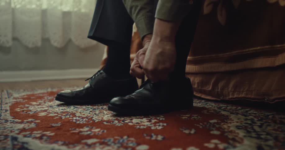 Man ties laces of black shoes while sitting on the edge of a bed over patterned rug