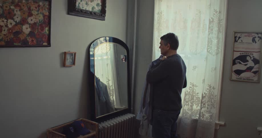 A man stands by a window holding up buttondown shirts on a hanger looking at them in front of a mirror