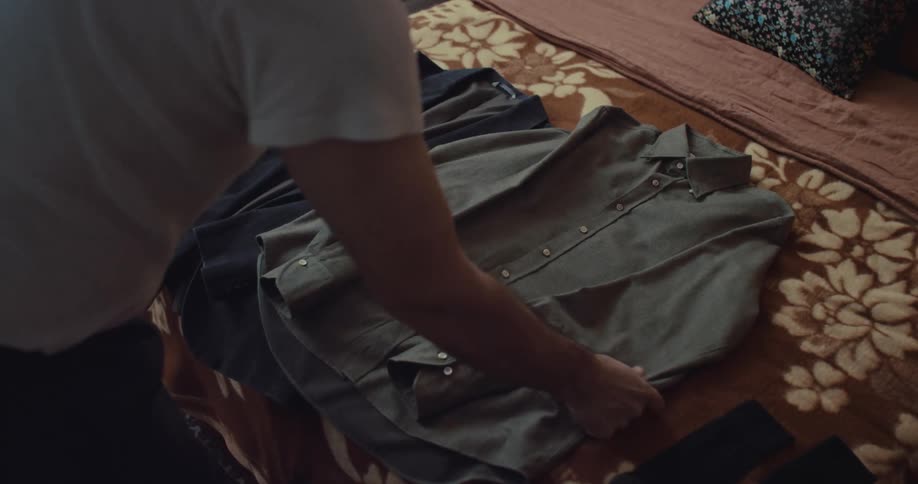 A man in a white tshirt carefully folds an olive green button down shirt on a bed covered with a brown floral patterned blanket next to a pair of dark pants