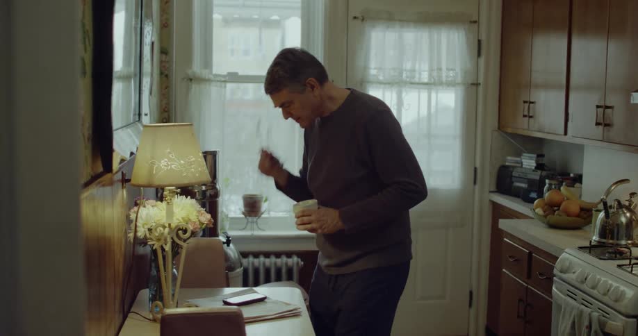 A middleaged man with short gray hair and glasses stands beside a small table in a softly lit kitchen holding a smartphone in one hand and a mug in the other while looking down at the screen then he pours some tea from a tea pot