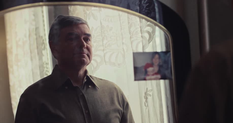An older man stands in front of a mirror in a softly lit room carefully fastening the top button of his shirt while looking at his reflection with a lace curtain behind him and a small photo of a child and woman attached to the mirror