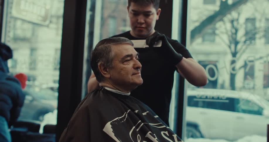 Barber cuts hair of man seated in chair in front of barbershop window