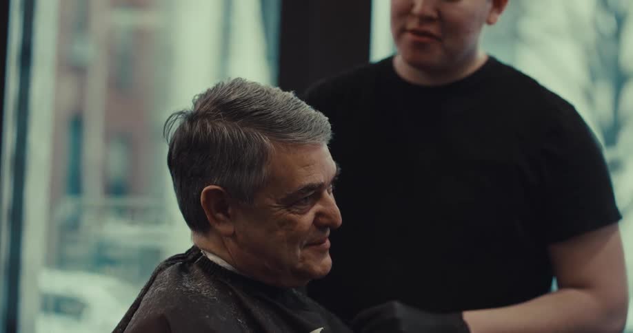 A barber wearing black gloves carefully trims the gray hair of an older man seated in a salon chair while the customer smiles slightly as he looks forward in the softly lit barbershop