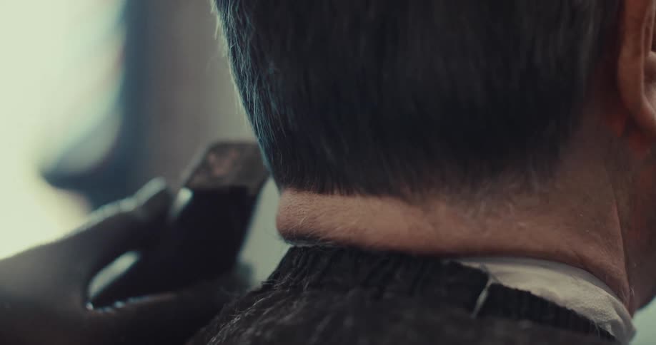 A barber wearing black gloves uses an electric clipper to trim the back of a mans neck during a professional haircut