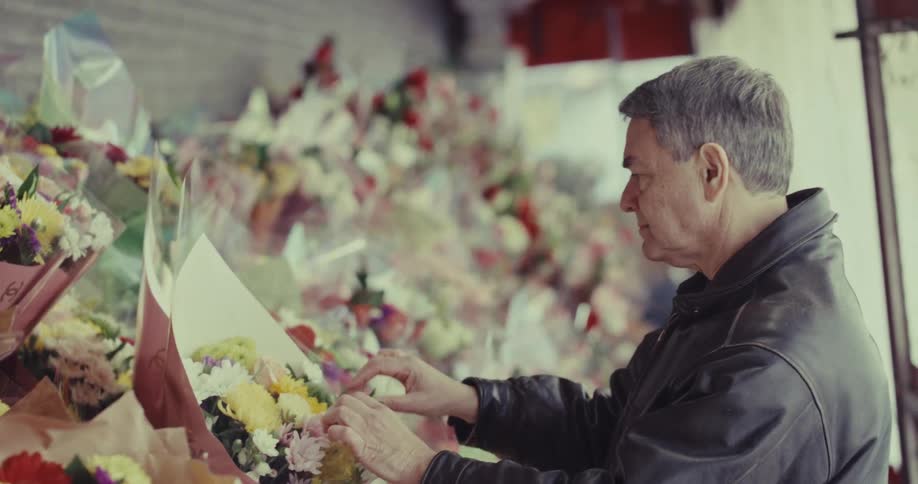 An older man wearing a dark leather jacket stands in a floral shop while carefully examining and touching a colorful bouquet of yellow and white flowers among many other wrapped arrangements on display