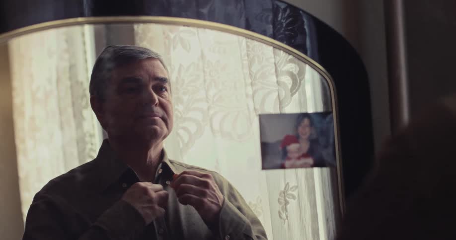 Man buttons shirt while looking at his reflection in mirror with photograph attached to the edge