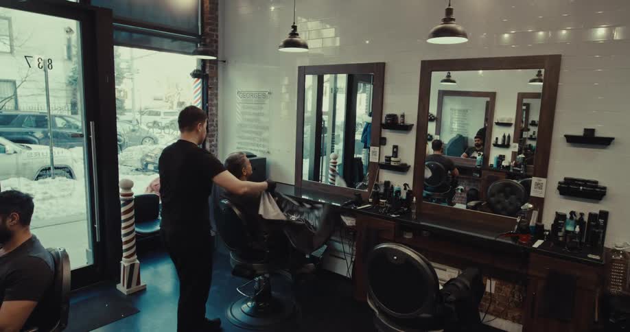 A barber grooming a customer in a modern barbershop with patrons nearby