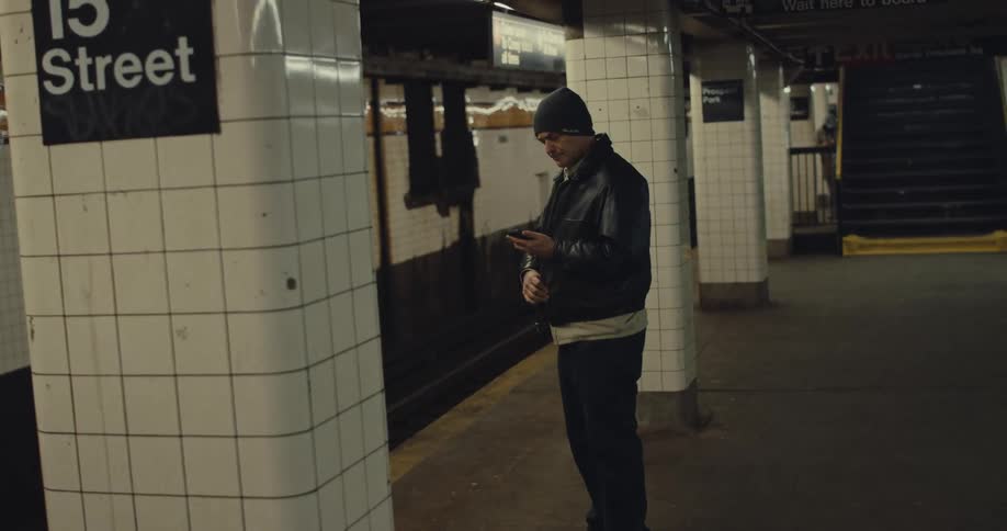 A man wearing a black leather jacket and beanie stands on a subway platform while checking his phone and looking down the tracks