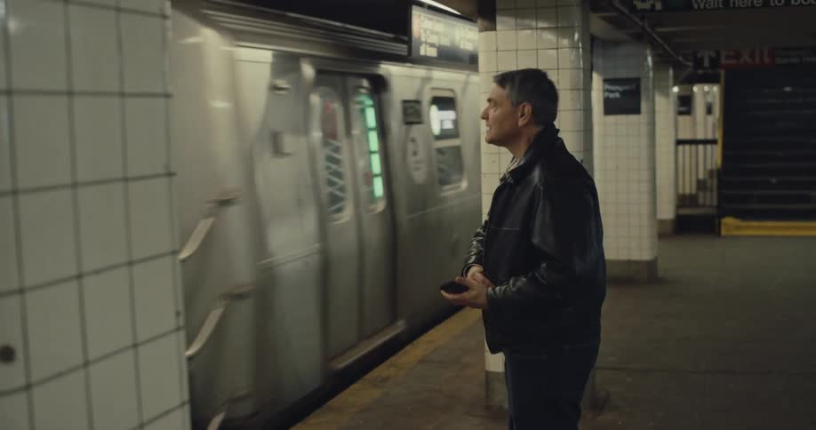 A man in a black jacket stands on a tiled subway platform watching a passing train as he checks his smart phone