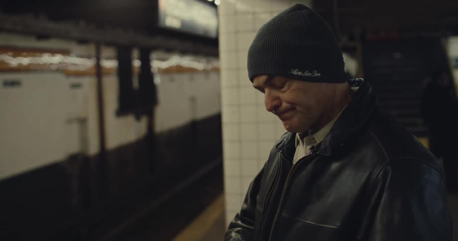 A man wearing a black leather jacket and a beanie stands on a subway platform waiting for a train