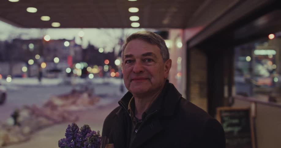 A middle aged man standing with a bouquet of flowers outside a storefront at dusk