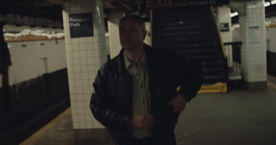 An older man in a leather jacket checks his phone while standing on a subway platformhe then tucks the phone into his pocket and begins walking toward the camera