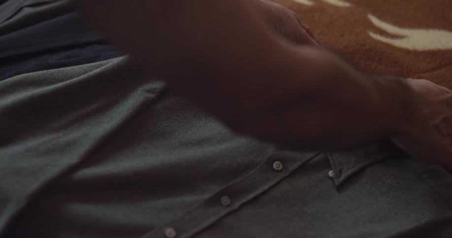 A pair of hands gently smooths and straightens a neatly folded buttondown shirt laid out on a patterned bedspread as if carefully preparing the clothing for wearing or packing