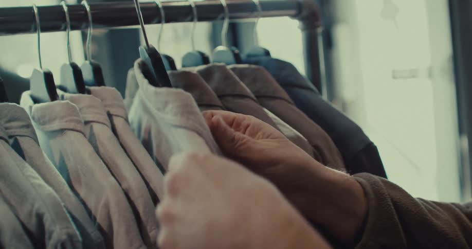 A mans hands move across a metal rack in a store carefully feeling the fabric of several hanging shirts before lifting a light colored button down to examine the collar and tag more closely