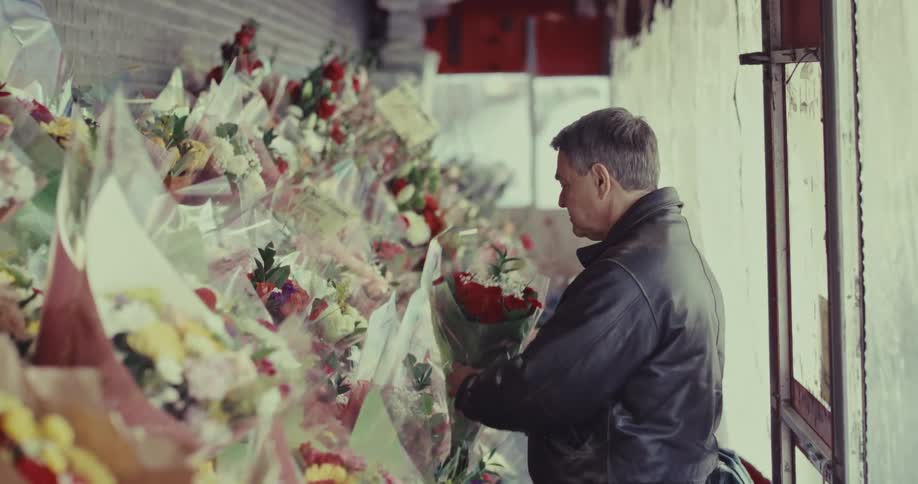 The video shows a middleaged man in a black leather jacket browsing through a dense outdoor display of floral bouquets he carefully examines a bunch of red roses before leaning down to look at other arrangements lined up along a brick wall