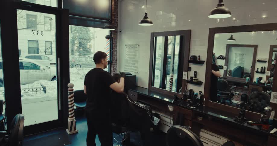 Barber cuts clients hair in chair inside barbershop with mirrors and streetfacing windows