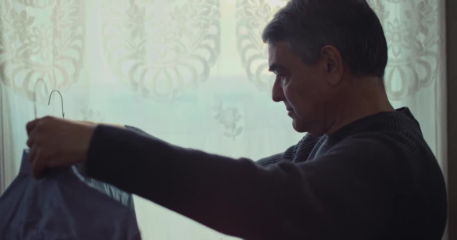 Man holds hanger with shirt while looking at his reflection beside curtained window