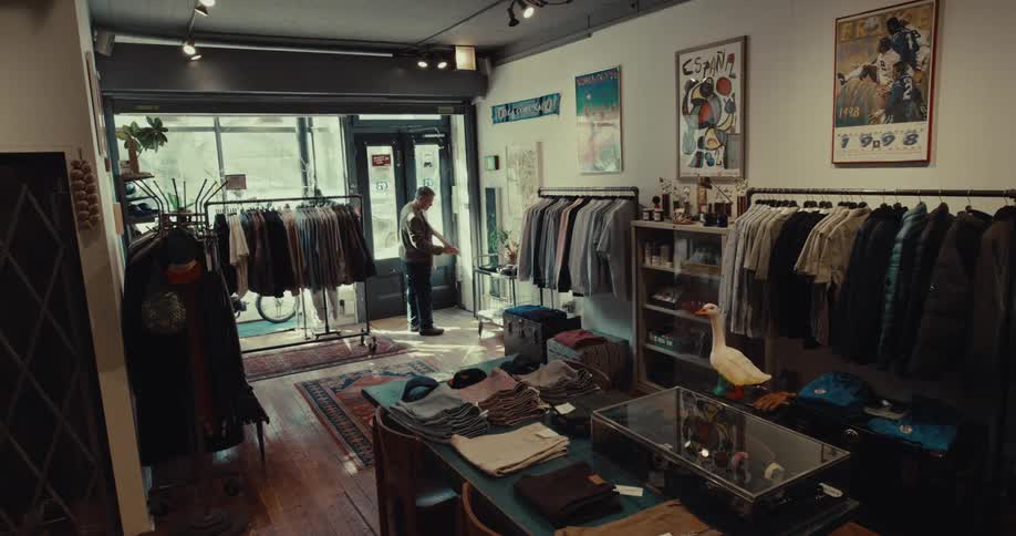 A man in a brown jacket walks toward the back of a small clothing boutique filled with vintage posters wooden floors and racks of apparel while a glass display case with a white goose statue sits in the foreground of the shop