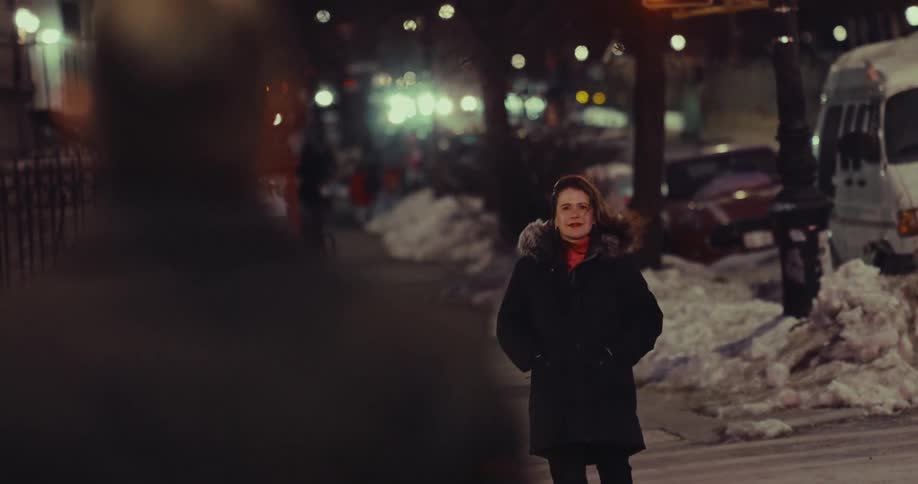 A woman in a winter coat stands on a snowy city sidewalk at night and smiles upon seeing someone off camera