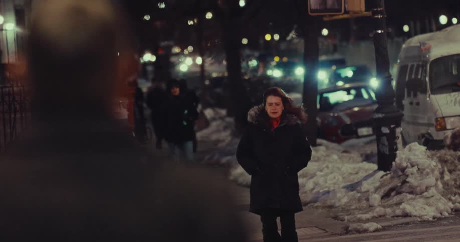 Woman walks along urban sidewalk at night between piled snow and parked cars