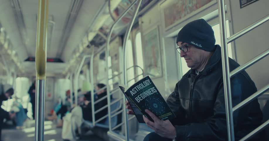 A man wearing a black beanie and leather jacket sits in a moving subway car while reading a book titled astrology for beginners