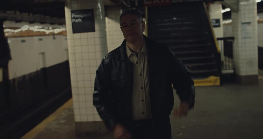 A man in a leather jacket walks down the stairs and onto a subway platform at prospect park station he reaches into his jacket pocket to retrieve his phone and checks it while continuing to walk