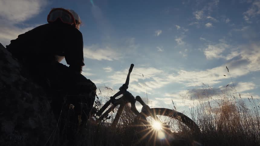 A silhouetted mountain biker sitting on a rock overlooking a grassy field at sunset