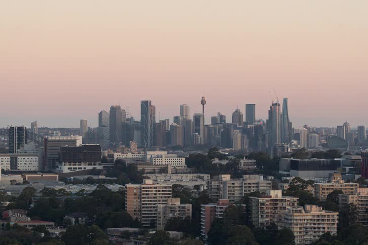 A time lapse view shows a wide city skyline with a tall tower as the natural daylight fades into a soft purple dusk and bright white and yellow lights begin to sparkle across the urban landscape and office buildings