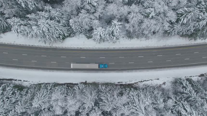 Straight down descent over a semi truck with a bright blue cab hauling a white trailer along a snowy interstate highway