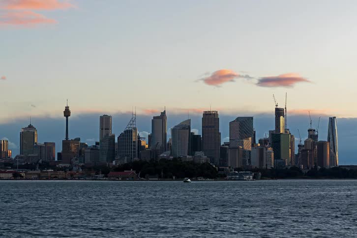 City skyline transitions from day to night as skyscrapers illuminate above the harbor while ferries and a cruise ship pass across the choppy blue water under a darkening sky filled with moving clouds