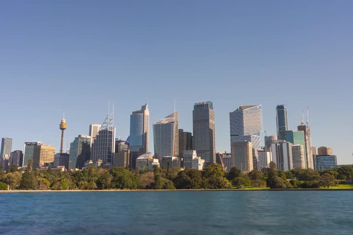 The video shows a wideangle static shot of the sydney skyline under a clear bright blue skythe citys highrise buildings and the sydney tower eye are visible across the harbor framed by green parkland and calm water in the foreground