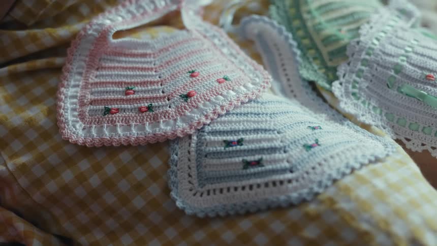 Handmade crocheted baby bibs with floral patterns are displayed on a yellow gingham fabric surface