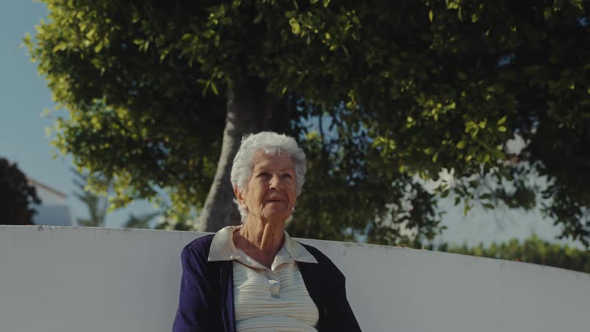 An elderly woman with short curly white hair sits outdoors on a bright day she is wearing a dark blue cardigan over a striped shirt and appears to be looking off into the distance
