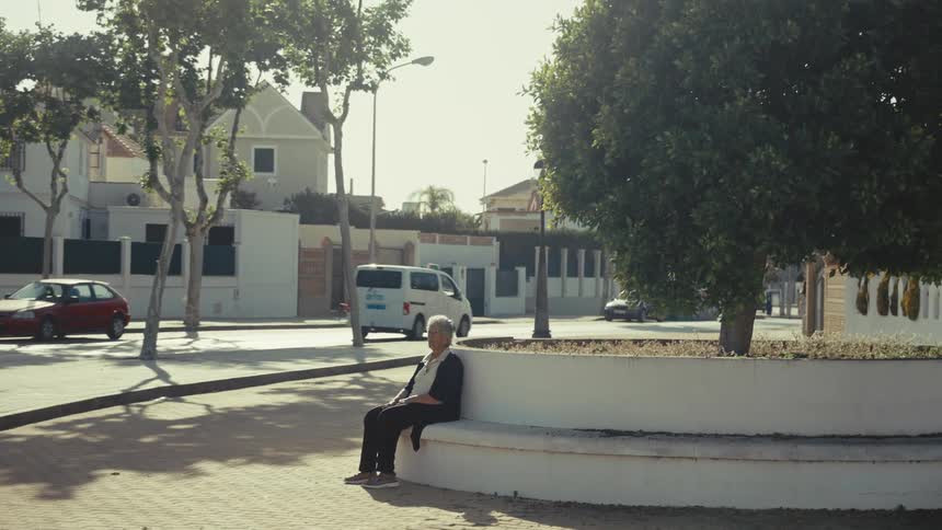 An elderly woman sits alone on a low white wall in a quiet suburban neighborhood during the daytime