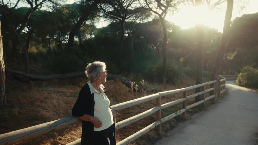 Elderly woman leans against a wooden fence looking around a single lane in the countryside during the daytime