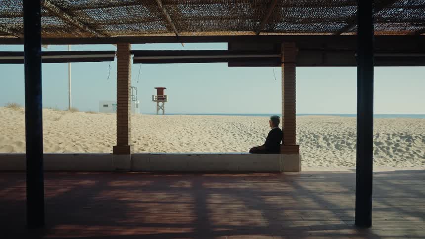 A woman sitting alone on a low white wall under a shaded beach structure gazing at the vast sandy beach