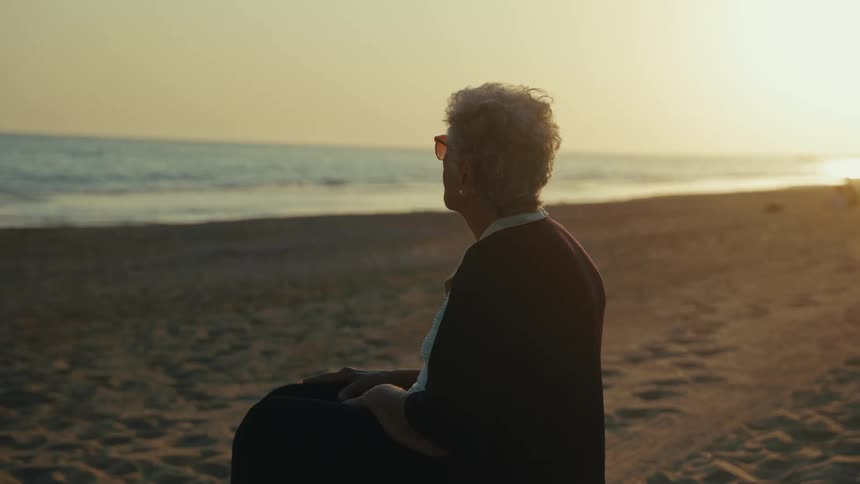 An elderly woman sits on a sandy beach at sunset gazing out toward the ocean wavesshe eventually removes her sunglasses to take in the view as the golden light fades over the horizon