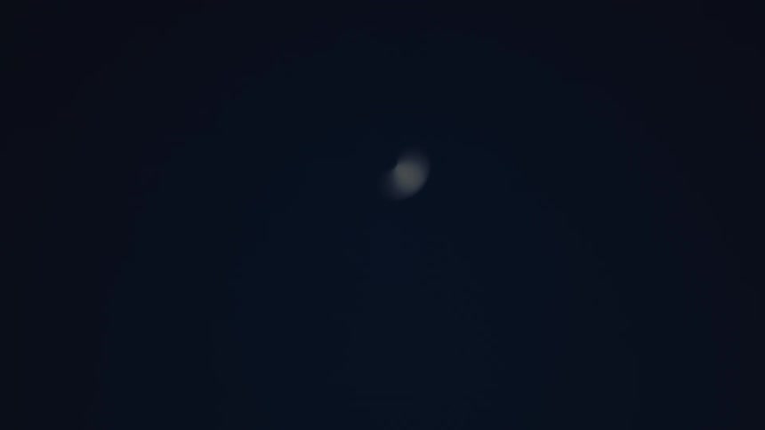 A thin bright crescent moon appearing and then fading against a dark nearblack sky a faint blurry circular light is visible initially before the sharp curve of the moon briefly comes into focus