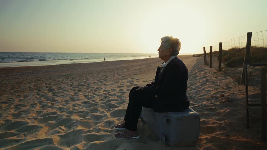An elderly woman sits alone on a bench by the beach gazing at the ocean during sunset