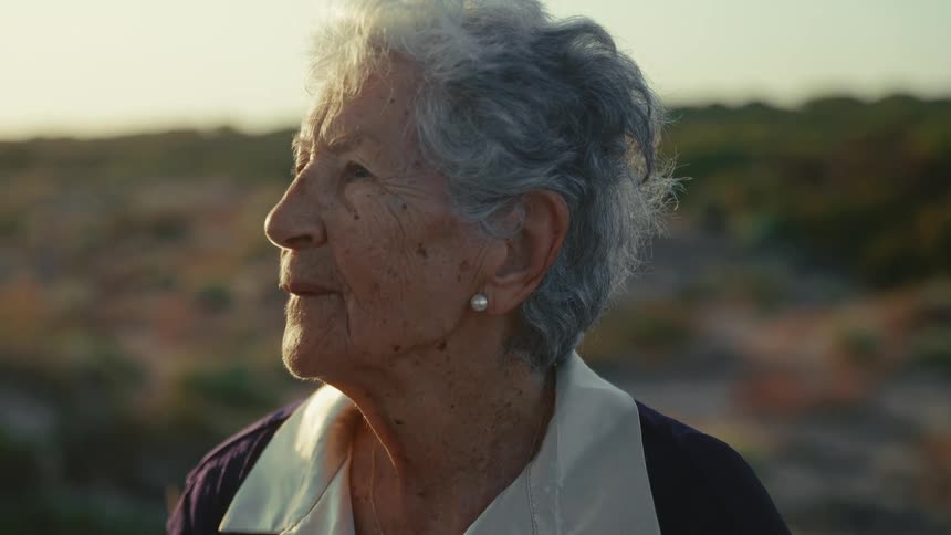 Elderly woman standing outdoors near a hill on a sunny day looks around