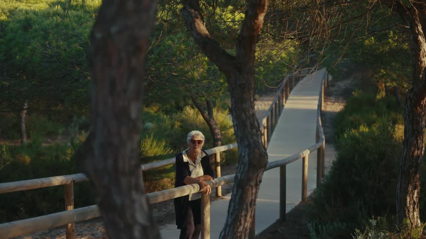 An elderly woman puts on her sunglasses while standing on a boardwalk in a forest