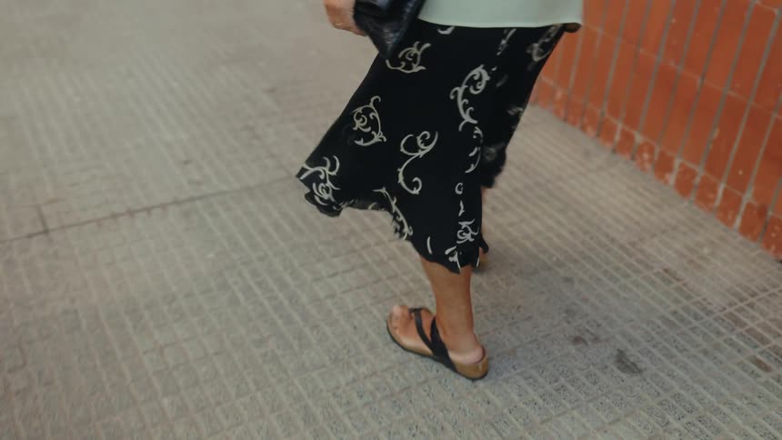 Elderly person walking beside a brick wall with a shoulder bag across the body