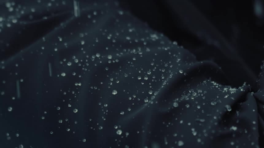 Close up shot of rain falling onto a dark black waterproof jacket where clear water droplets bead and roll across the textured synthetic fabric surface during a heavy downpour in a moody outdoor environment