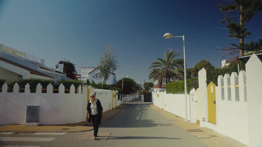 Person walking along a residential street between white fences under a blue sky