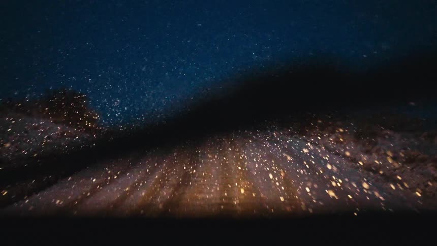 The perspective of driving down a snowcovered road at night where the vehicles headlights illuminate thick snowflakes falling against a dark blue skythe two lines of tire tracks create a strong sense of movement carving a path through the dusting o