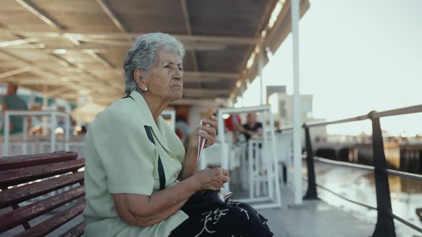 Smiling elderly woman eats dessert while sitting on a bench in a waterfront pavilion during the daytime