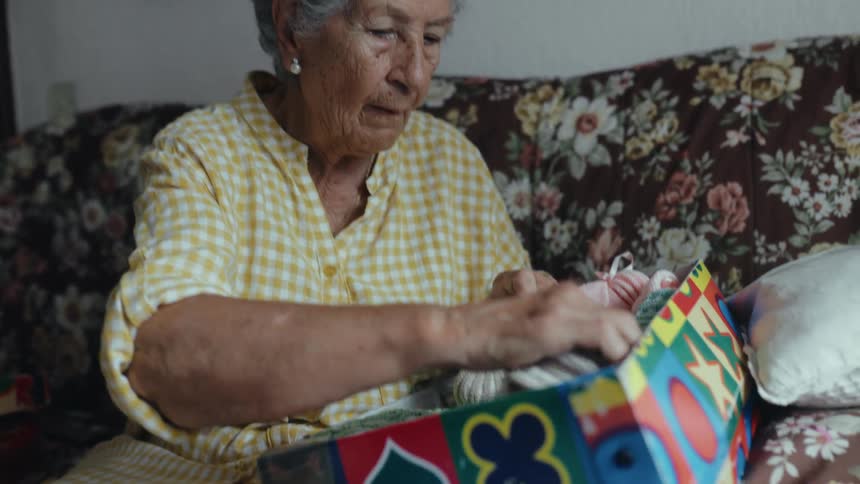 Elderly woman removes baby clothes from a box while sitting on the couch in her living room during the daytime